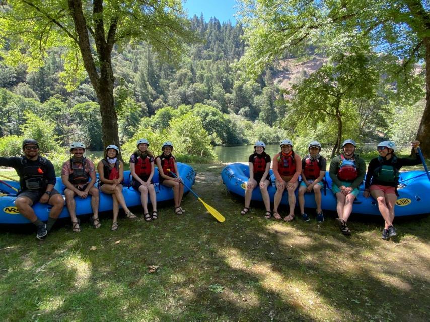 A group of peak adventures whitewater rafters sitting on the edge of a raft posing for a photo on the banks of on the south fork of the american river.